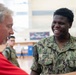 A U.S. Navy sailor named Brown is smiling broadly while interacting with an older civilian man in a red shirt, appearing to shake hands or engage in conversation. The setting appears to be an indoor gymnasium or recreational facility. Additional military personnel in camouflage uniforms are visible in the background.
