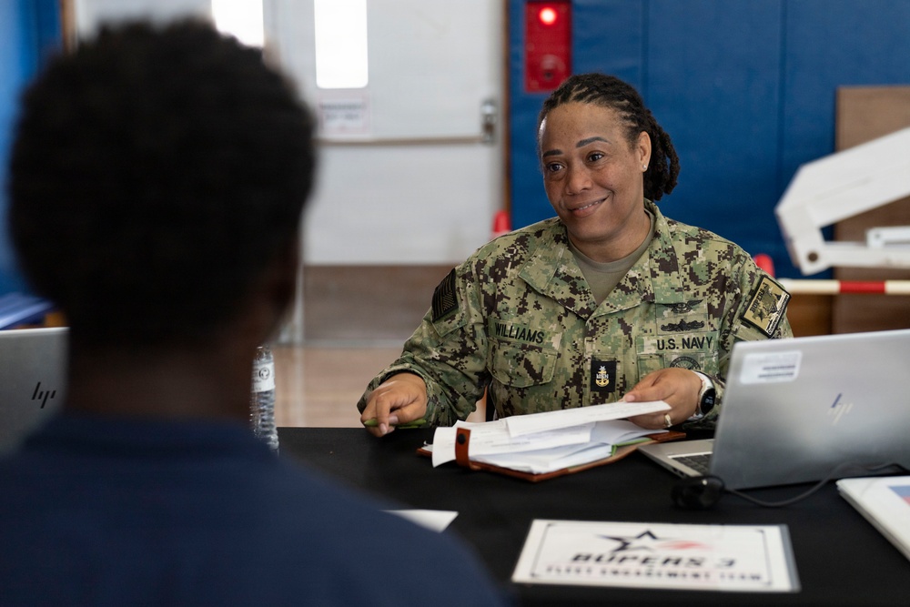 A U.S. Navy service member named Williams is seated at a table conducting what appears to be an administrative consultation or briefing session. She is in NWU (Navy Working Uniform) camouflage and is engaged with another individual seated across from her. A placard on the table reads 'BUPERS 3 Fleet Engagement Team,' indicating this is a Navy Bureau of Personnel outreach event.