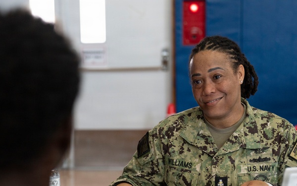 A U.S. Navy service member named Williams is seated at a table conducting what appears to be an administrative consultation or briefing session. She is in NWU (Navy Working Uniform) camouflage and is engaged with another individual seated across from her. A placard on the table reads 'BUPERS 3 Fleet Engagement Team,' indicating this is a Navy Bureau of Personnel outreach event.
