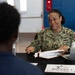 A U.S. Navy service member named Williams is seated at a table conducting what appears to be an administrative consultation or briefing session. She is in NWU (Navy Working Uniform) camouflage and is engaged with another individual seated across from her. A placard on the table reads 'BUPERS 3 Fleet Engagement Team,' indicating this is a Navy Bureau of Personnel outreach event.