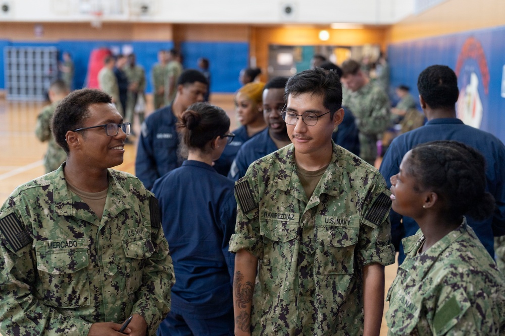 A group of U.S. Navy personnel in NWU Type I (digital camouflage) and Navy Working Uniform (blue) are gathered inside what appears to be a military gymnasium or recreational facility. Three sailors are prominently visible in the foreground, with name tapes reading 'MERCADO' and 'RAMREZDIAZ' on their uniforms. The setting suggests a formation, briefing, or social gathering event.