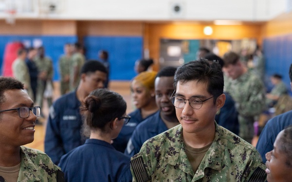 A group of U.S. Navy personnel in NWU Type I (digital camouflage) and Navy Working Uniform (blue) are gathered inside what appears to be a military gymnasium or recreational facility. Three sailors are prominently visible in the foreground, with name tapes reading 'MERCADO' and 'RAMREZDIAZ' on their uniforms. The setting suggests a formation, briefing, or social gathering event.