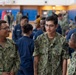 A group of U.S. Navy personnel in NWU Type I (digital camouflage) and Navy Working Uniform (blue) are gathered inside what appears to be a military gymnasium or recreational facility. Three sailors are prominently visible in the foreground, with name tapes reading 'MERCADO' and 'RAMREZDIAZ' on their uniforms. The setting suggests a formation, briefing, or social gathering event.