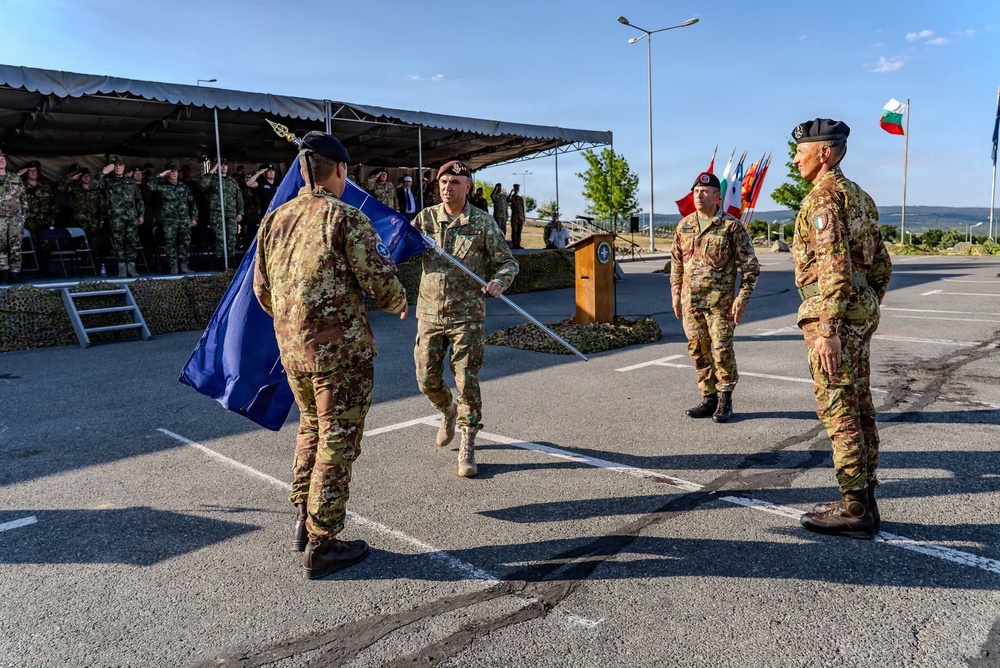 A military ceremony is taking place on a paved parade ground, likely a change of command or unit activation/deactivation ceremony. Several soldiers in camouflage uniforms are participating in a flag transfer ritual, with a blue flag (appearing to be a NATO flag) being passed between personnel. A formation of troops stands at attention under a tent canopy in the background, with multiple national flags visible including what appears to be the Bulgarian flag.