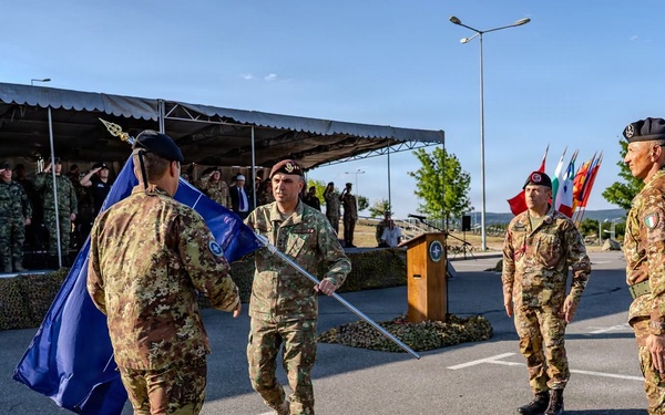 A military ceremony is taking place on a paved parade ground, likely a change of command or unit activation/deactivation ceremony. Several soldiers in camouflage uniforms are participating in a flag transfer ritual, with a blue flag (appearing to be a NATO flag) being passed between personnel. A formation of troops stands at attention under a tent canopy in the background, with multiple national flags visible including what appears to be the Bulgarian flag.