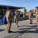 A military ceremony is taking place on a paved parade ground, likely a change of command or unit activation/deactivation ceremony. Several soldiers in camouflage uniforms are participating in a flag transfer ritual, with a blue flag (appearing to be a NATO flag) being passed between personnel. A formation of troops stands at attention under a tent canopy in the background, with multiple national flags visible including what appears to be the Bulgarian flag.