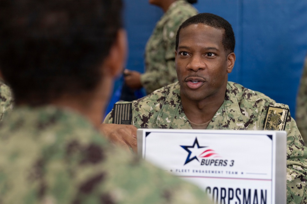 A U.S. Navy service member in AOR2 camouflage uniform is engaged in a conversation at a BUPERS 3 Fleet Engagement Team table. A sign on the table indicates the role of 'Corpsman,' suggesting this is a career counseling or administrative engagement event. The setting appears to be an indoor military facility with blue padded walls visible in the background.