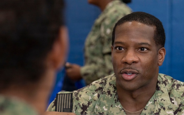 A U.S. Navy service member in AOR2 camouflage uniform is engaged in a conversation at a BUPERS 3 Fleet Engagement Team table. A sign on the table indicates the role of 'Corpsman,' suggesting this is a career counseling or administrative engagement event. The setting appears to be an indoor military facility with blue padded walls visible in the background.