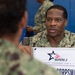 A U.S. Navy service member in AOR2 camouflage uniform is engaged in a conversation at a BUPERS 3 Fleet Engagement Team table. A sign on the table indicates the role of 'Corpsman,' suggesting this is a career counseling or administrative engagement event. The setting appears to be an indoor military facility with blue padded walls visible in the background.