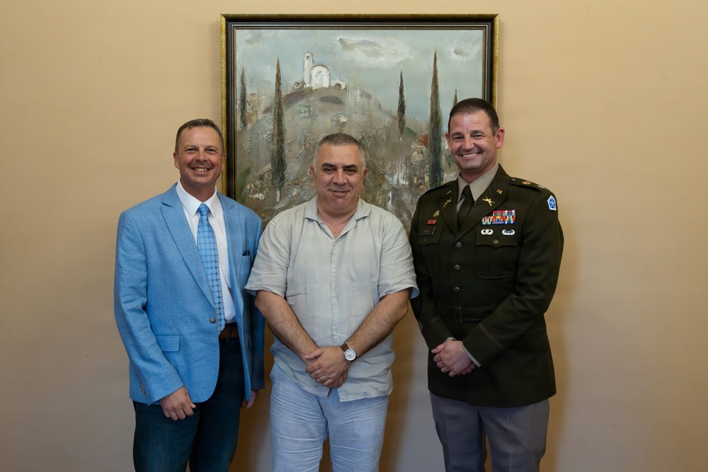 Three men are posing together for a formal photograph in an indoor setting. One man is wearing a U.S. Army dress uniform with visible rank insignia, medals, and unit patches, while the other two are in civilian attire. They are standing in front of a landscape painting on a beige wall.