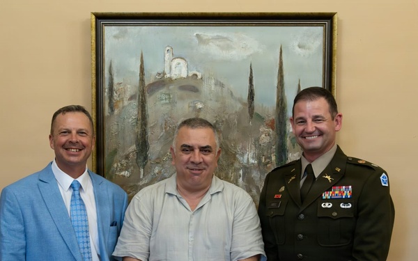 Three men are posing together for a formal photograph in an indoor setting. One man is wearing a U.S. Army dress uniform with visible rank insignia, medals, and unit patches, while the other two are in civilian attire. They are standing in front of a landscape painting on a beige wall.