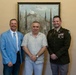 Three men are posing together for a formal photograph in an indoor setting. One man is wearing a U.S. Army dress uniform with visible rank insignia, medals, and unit patches, while the other two are in civilian attire. They are standing in front of a landscape painting on a beige wall.