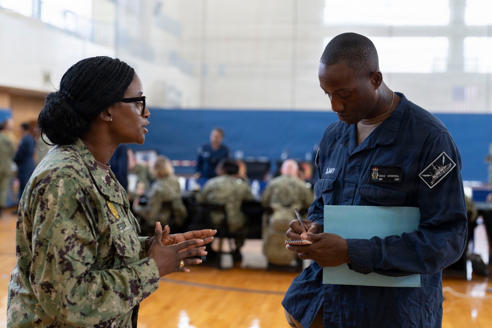 Two U.S. Navy personnel are engaged in a conversation inside what appears to be a military gymnasium or community center. The individual on the left is wearing Army Combat Uniform (ACU) pattern camouflage, while the person on the right is in Navy Working Uniform (NWU) blue coveralls and is taking notes on a small notepad while holding a blue folder. In the background, multiple other uniformed personnel are seated at tables, suggesting an organized military event or briefing.