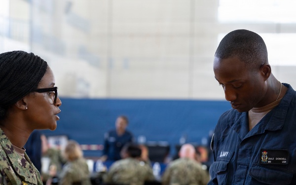 Two U.S. Navy personnel are engaged in a conversation inside what appears to be a military gymnasium or community center. The individual on the left is wearing Army Combat Uniform (ACU) pattern camouflage, while the person on the right is in Navy Working Uniform (NWU) blue coveralls and is taking notes on a small notepad while holding a blue folder. In the background, multiple other uniformed personnel are seated at tables, suggesting an organized military event or briefing.