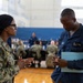 Two U.S. Navy personnel are engaged in a conversation inside what appears to be a military gymnasium or community center. The individual on the left is wearing Army Combat Uniform (ACU) pattern camouflage, while the person on the right is in Navy Working Uniform (NWU) blue coveralls and is taking notes on a small notepad while holding a blue folder. In the background, multiple other uniformed personnel are seated at tables, suggesting an organized military event or briefing.