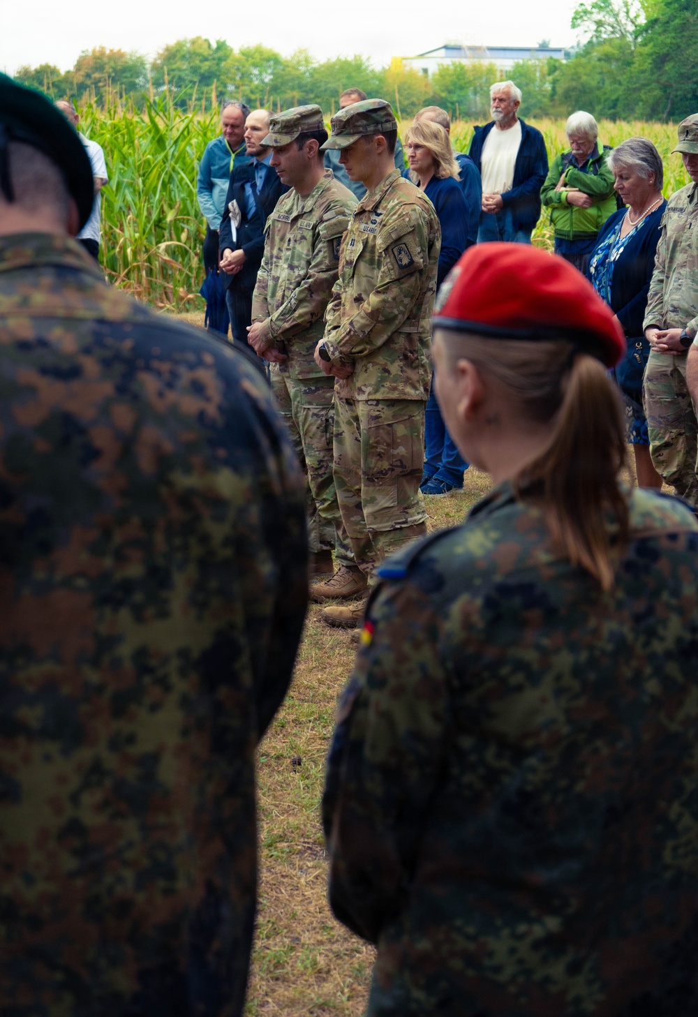 A mixed military and civilian gathering is taking place outdoors near a cornfield, appearing to be a solemn ceremony or memorial event. Personnel in U.S. Army OCP (Operational Camouflage Pattern) uniforms stand alongside individuals in German Bundeswehr Flecktarn camouflage uniforms, suggesting a joint multinational military event. Civilians in casual and semi-formal attire are also present in the background, with heads bowed in what appears to be a moment of reflection or prayer.