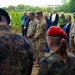 A mixed military and civilian gathering is taking place outdoors near a cornfield, appearing to be a solemn ceremony or memorial event. Personnel in U.S. Army OCP (Operational Camouflage Pattern) uniforms stand alongside individuals in German Bundeswehr Flecktarn camouflage uniforms, suggesting a joint multinational military event. Civilians in casual and semi-formal attire are also present in the background, with heads bowed in what appears to be a moment of reflection or prayer.