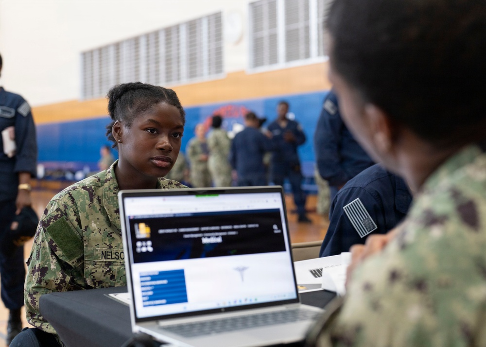 A U.S. Navy service member with the name tag 'NELSON' is seated at a table with an open laptop displaying what appears to be a medical-related webpage, engaged in conversation with another service member across the table. The setting appears to be a gymnasium or large indoor facility being used for a military administrative or processing event. Multiple uniformed personnel are visible in the background.
