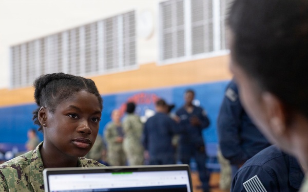 A U.S. Navy service member with the name tag 'NELSON' is seated at a table with an open laptop displaying what appears to be a medical-related webpage, engaged in conversation with another service member across the table. The setting appears to be a gymnasium or large indoor facility being used for a military administrative or processing event. Multiple uniformed personnel are visible in the background.