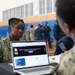 A U.S. Navy service member with the name tag 'NELSON' is seated at a table with an open laptop displaying what appears to be a medical-related webpage, engaged in conversation with another service member across the table. The setting appears to be a gymnasium or large indoor facility being used for a military administrative or processing event. Multiple uniformed personnel are visible in the background.