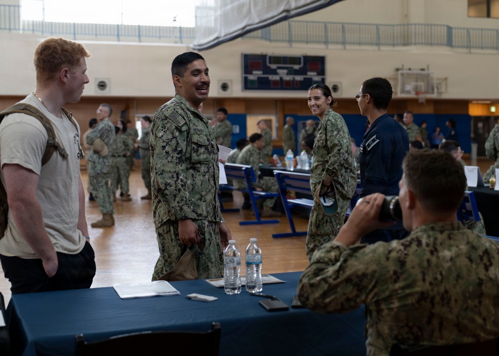 Military personnel, appearing to be U.S. Navy sailors based on their NWU (Navy Working Uniform) camouflage pattern, are gathered inside a gymnasium for what appears to be an administrative processing or check-in event. Multiple tables are set up with paperwork and water bottles, and personnel are engaged in conversations and processing activities. The setting resembles a mobilization, deployment processing, or administrative muster event.