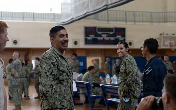 Military personnel, appearing to be U.S. Navy sailors based on their NWU (Navy Working Uniform) camouflage pattern, are gathered inside a gymnasium for what appears to be an administrative processing or check-in event. Multiple tables are set up with paperwork and water bottles, and personnel are engaged in conversations and processing activities. The setting resembles a mobilization, deployment processing, or administrative muster event.