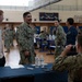 Military personnel, appearing to be U.S. Navy sailors based on their NWU (Navy Working Uniform) camouflage pattern, are gathered inside a gymnasium for what appears to be an administrative processing or check-in event. Multiple tables are set up with paperwork and water bottles, and personnel are engaged in conversations and processing activities. The setting resembles a mobilization, deployment processing, or administrative muster event.