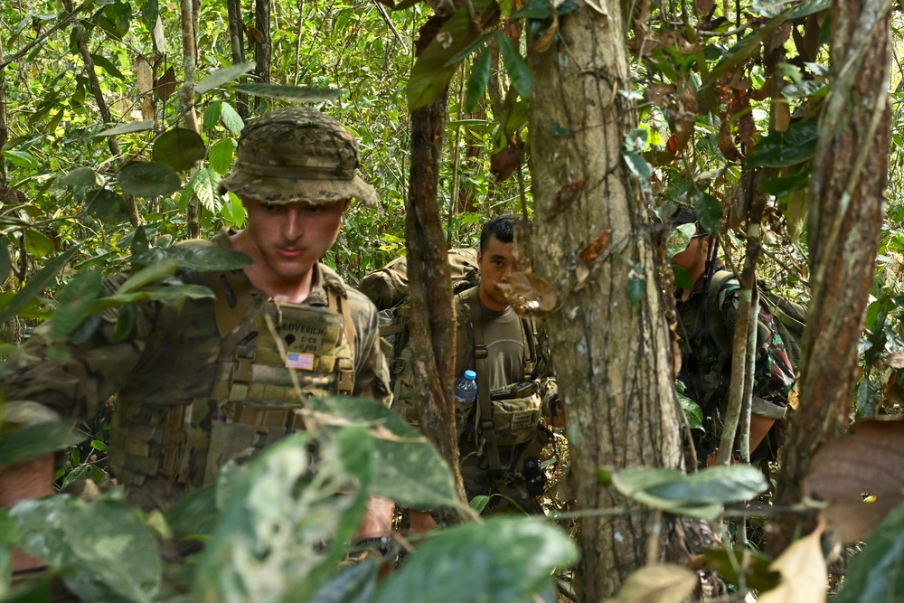 A group of military personnel are navigating through dense jungle terrain. At least three soldiers are visible, with the foremost wearing US Army multicam uniform with an American flag patch and a name tape reading 'COVERICH'. The soldiers appear to be on a patrol or training exercise in a tropical forest environment.