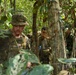 A group of military personnel are navigating through dense jungle terrain. At least three soldiers are visible, with the foremost wearing US Army multicam uniform with an American flag patch and a name tape reading 'COVERICH'. The soldiers appear to be on a patrol or training exercise in a tropical forest environment.