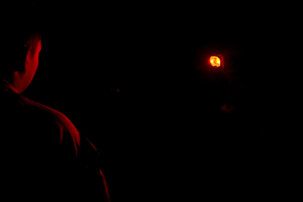 A very dark image showing a person illuminated by red light on the left side, with a small orange/amber light source visible in the upper right area. The scene appears to be taken in near-total darkness, possibly outdoors at night. A second partially visible figure or hand can be faintly detected near the right light source.