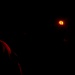 A very dark image showing a person illuminated by red light on the left side, with a small orange/amber light source visible in the upper right area. The scene appears to be taken in near-total darkness, possibly outdoors at night. A second partially visible figure or hand can be faintly detected near the right light source.