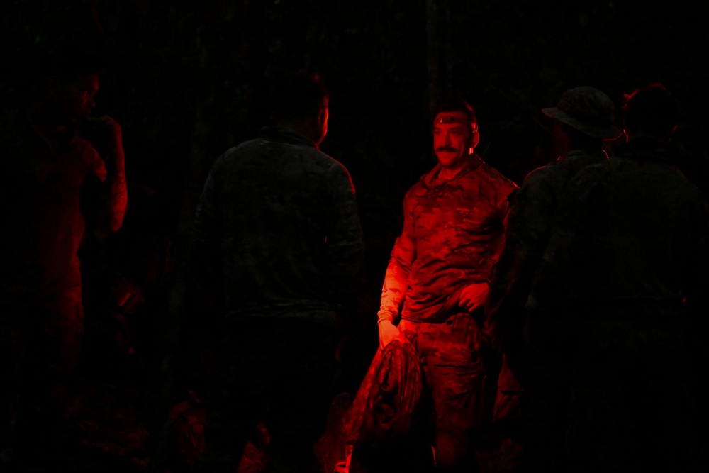 A group of individuals in military camouflage uniforms are gathered in a dark environment, illuminated primarily by a red light source. The central figure, a man with a mustache, is most clearly visible as the red light falls directly on him. The scene appears to depict a nighttime military briefing, rest period, or assembly.