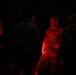 A group of individuals in military camouflage uniforms are gathered in a dark environment, illuminated primarily by a red light source. The central figure, a man with a mustache, is most clearly visible as the red light falls directly on him. The scene appears to depict a nighttime military briefing, rest period, or assembly.