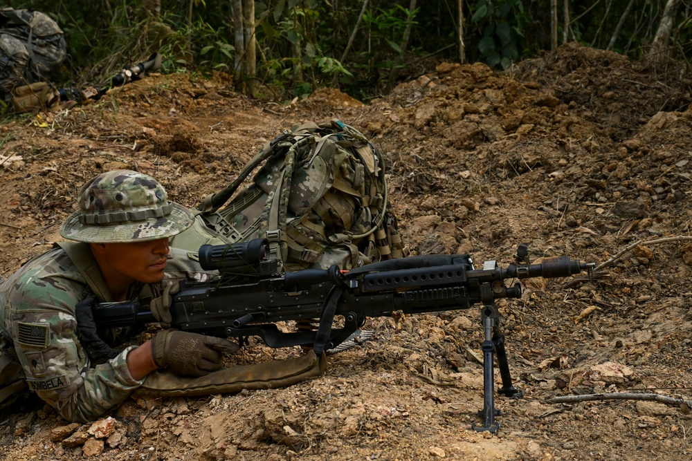 A U.S. Army soldier in MultiCam/OCP camouflage is in a prone firing position in a jungle or forested training environment. The soldier is operating what appears to be an M249 SAW (Squad Automatic Weapon) or similar light machine gun mounted on a bipod. A tactical backpack is positioned nearby, and at least one additional soldier is visible in the background.