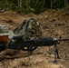 A U.S. Army soldier in MultiCam/OCP camouflage is in a prone firing position in a jungle or forested training environment. The soldier is operating what appears to be an M249 SAW (Squad Automatic Weapon) or similar light machine gun mounted on a bipod. A tactical backpack is positioned nearby, and at least one additional soldier is visible in the background.