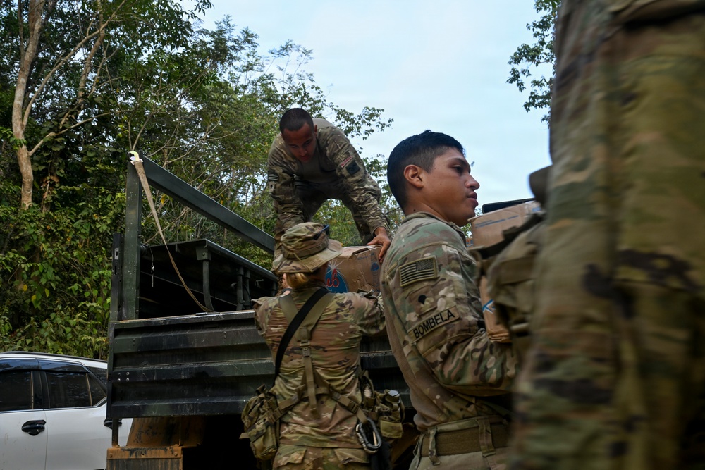 U.S. Army soldiers in MultiCam/OCP camouflage uniforms are unloading or loading boxes from a military truck in a tropical or jungle environment. At least one soldier is identifiable by the name tape 'BOMBELA' on their uniform, and all personnel display U.S. flag patches. The operation appears to be a logistics or humanitarian aid distribution mission.