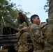U.S. Army soldiers in MultiCam/OCP camouflage uniforms are unloading or loading boxes from a military truck in a tropical or jungle environment. At least one soldier is identifiable by the name tape 'BOMBELA' on their uniform, and all personnel display U.S. flag patches. The operation appears to be a logistics or humanitarian aid distribution mission.