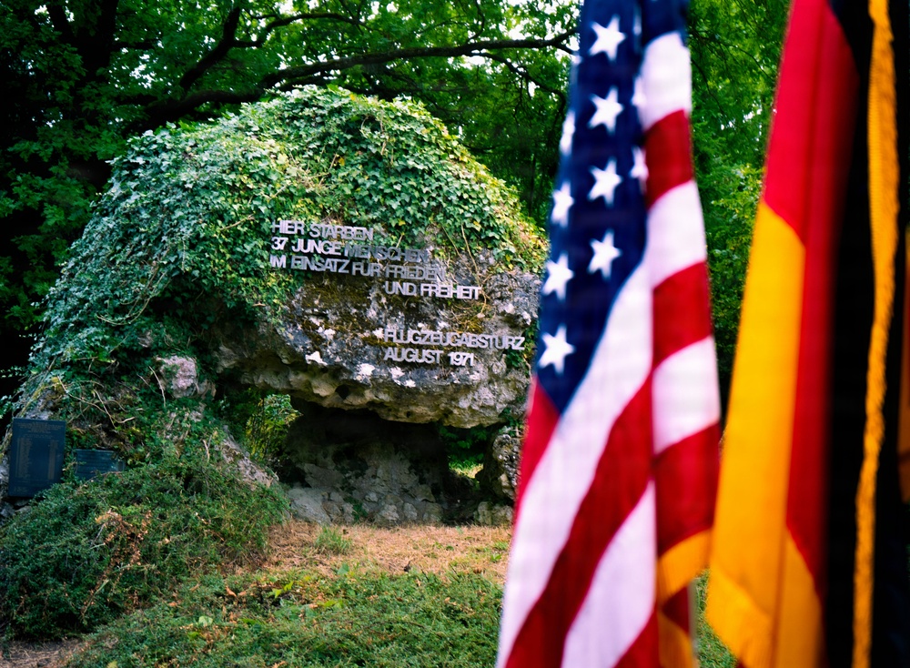 A memorial stone covered in ivy bears a German-language inscription commemorating 37 young people who died in a plane crash (Flugzeugabsturz) in August 1971. The monument is flanked by an American flag on the left and a German flag on the right, indicating a joint commemoration. The setting is an outdoor wooded area with lush green vegetation surrounding the large boulder-like memorial.
