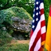A memorial stone covered in ivy bears a German-language inscription commemorating 37 young people who died in a plane crash (Flugzeugabsturz) in August 1971. The monument is flanked by an American flag on the left and a German flag on the right, indicating a joint commemoration. The setting is an outdoor wooded area with lush green vegetation surrounding the large boulder-like memorial.