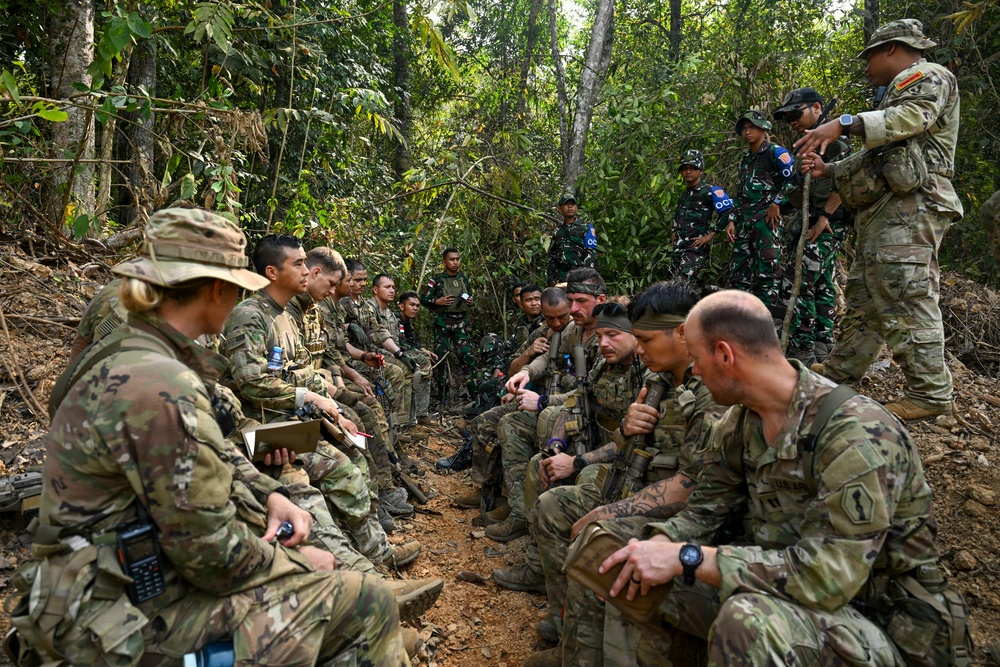 A group of military personnel from at least two different nations are gathered in a jungle/forest environment, appearing to conduct a briefing or after-action review. Some soldiers wear MultiCam/OCP uniforms consistent with US Army, while others wear distinctive camouflage patterns consistent with Indonesian Armed Forces (TNI). The setting and mixed composition suggest a joint military training exercise.