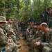 A group of military personnel from at least two different nations are gathered in a jungle/forest environment, appearing to conduct a briefing or after-action review. Some soldiers wear MultiCam/OCP uniforms consistent with US Army, while others wear distinctive camouflage patterns consistent with Indonesian Armed Forces (TNI). The setting and mixed composition suggest a joint military training exercise.