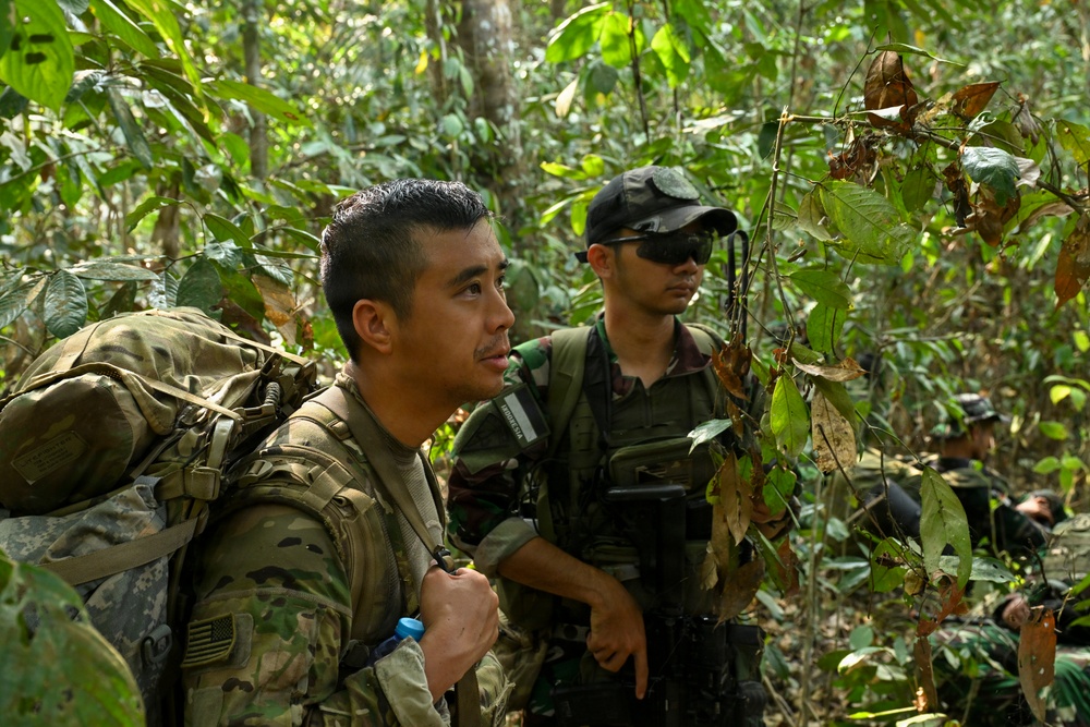 Two armed military personnel are moving through dense tropical jungle terrain. The foreground soldier wears US Army multicam uniform with a large rucksack and a visible US flag patch on the left shoulder. A second soldier wearing a black cap and tactical vest follows closely behind, with additional personnel partially visible in the background.