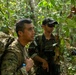Two armed military personnel are moving through dense tropical jungle terrain. The foreground soldier wears US Army multicam uniform with a large rucksack and a visible US flag patch on the left shoulder. A second soldier wearing a black cap and tactical vest follows closely behind, with additional personnel partially visible in the background.