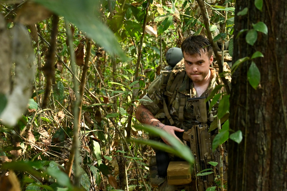A male soldier in camouflage military gear navigates through dense jungle or forest terrain. He is carrying a large tactical backpack and what appears to be a rifle, pushing through thick vegetation. His clothing and equipment suggest active military field operations or training exercise.