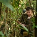 A male soldier in camouflage military gear navigates through dense jungle or forest terrain. He is carrying a large tactical backpack and what appears to be a rifle, pushing through thick vegetation. His clothing and equipment suggest active military field operations or training exercise.