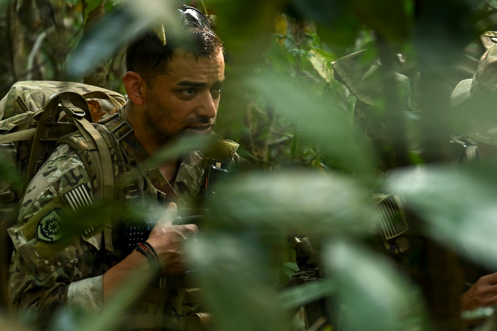 A U.S. military soldier in MultiCam/OCP camouflage uniform is navigating through dense jungle or tropical vegetation. The soldier is equipped with a tactical backpack, body armor, and appears to be holding a weapon or radio. A second soldier in similar uniform is partially visible to the right, suggesting a small unit patrol or training exercise.