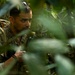 A U.S. military soldier in MultiCam/OCP camouflage uniform is navigating through dense jungle or tropical vegetation. The soldier is equipped with a tactical backpack, body armor, and appears to be holding a weapon or radio. A second soldier in similar uniform is partially visible to the right, suggesting a small unit patrol or training exercise.