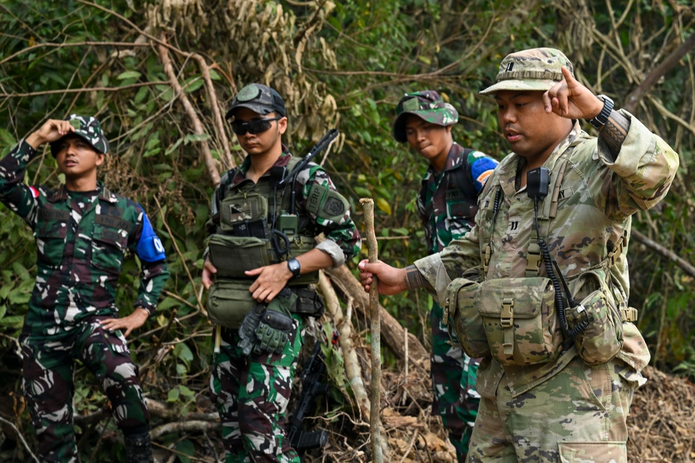 The image shows four military personnel in a jungle or forested environment, appearing to be engaged in a joint training exercise or patrol operation. Three individuals are wearing Indonesian Armed Forces (TNI) camouflage uniforms, while one person on the right wears a US Army MultiCam (OCP) uniform. The soldiers appear to be communicating or receiving instructions, with some gesturing toward their ears or heads.