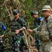 The image shows four military personnel in a jungle or forested environment, appearing to be engaged in a joint training exercise or patrol operation. Three individuals are wearing Indonesian Armed Forces (TNI) camouflage uniforms, while one person on the right wears a US Army MultiCam (OCP) uniform. The soldiers appear to be communicating or receiving instructions, with some gesturing toward their ears or heads.