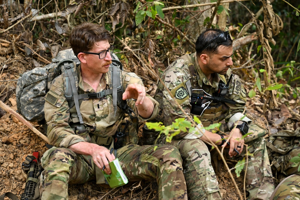 Two military personnel in MultiCam/OCP camouflage uniforms are seated on a hillside in a jungle or forested environment. The soldier on the left appears to be briefing or explaining something while holding a small notebook and gesturing with his hand. Both carry tactical gear including radios and backpacks, and wear US flag patches on their shoulders.